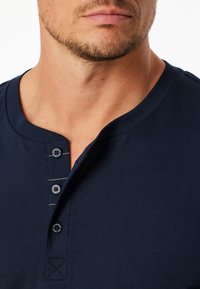 Námořnická modrá košile henley s třemi černými knoflíky a bílým švy na léze, vyrobená z měkké a hladké tkaniny.