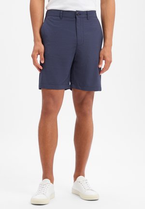 Mężczyzna w ciemnoniebieskich, teksturowanych szortach do kolan i białych sneakersach, stojący na białym tle.