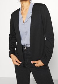 Černý, otevřený blazer s dlouhými rukávy, vyrobený z měkké látky. Nositelný přes světle modrou košili s knoflíky. Jednoduchý design, žádné viditelné vzory.