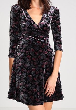 Femme portant une robe en velours noir avec un motif floral rose et gris, manches trois-quarts et un corsage cache-cœur avec décolleté en V.