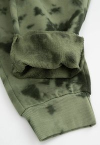 Kardigan w kolorze oliwkowym, z rękawem w technice tie-dye, o prążkowanej fakturze; miękki materiał; ma zaokrągloną krawędź i luźny krój, podkreślający wygodę.