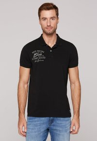 Polo nero con una texture sottile, dotato di una patta con tre bottoni, logo ricamato in grigio e una tasca sul petto. Abbinato a jeans blu.