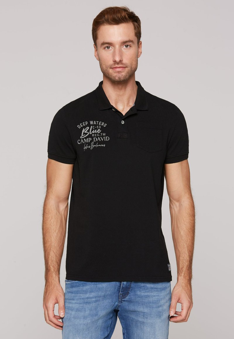 Polo nero con una texture sottile, dotato di una patta con tre bottoni, logo ricamato in grigio e una tasca sul petto. Abbinato a jeans blu.