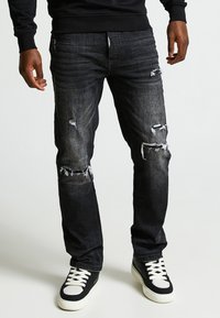 Vaqueros de mezclilla de lavado oscuro con un corte recto, que presentan detalles desgastados y desgarrones en las rodillas, combinados con unas zapatillas altas en blanco y negro.