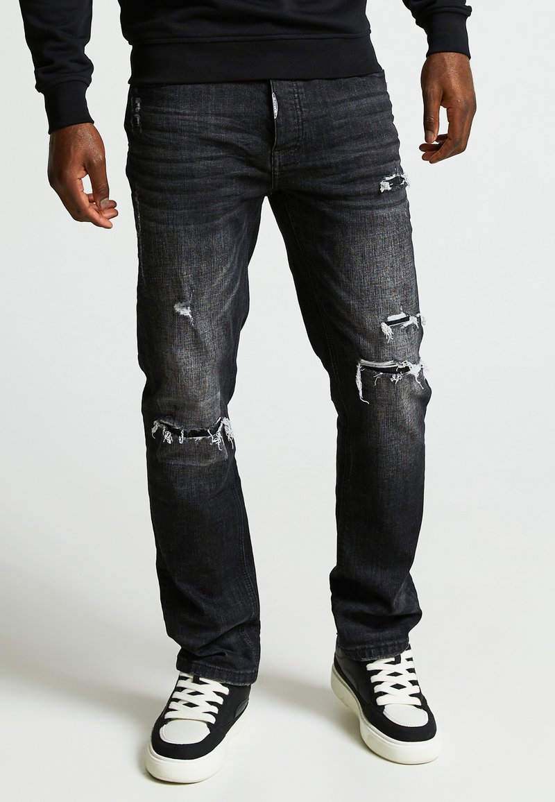 Vaqueros de mezclilla de lavado oscuro con un corte recto, que presentan detalles desgastados y desgarrones en las rodillas, combinados con unas zapatillas altas en blanco y negro.