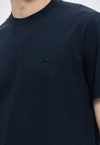 Homme portant une chemise bleu marine à manches courtes avec col côtelé et un petit logo crocodile brodé sur la poitrine gauche.