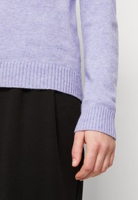 Mano reposando al lado del cuerpo, vistiendo un suéter de punto morado claro con pu�ños acanalados y una prenda negra debajo.