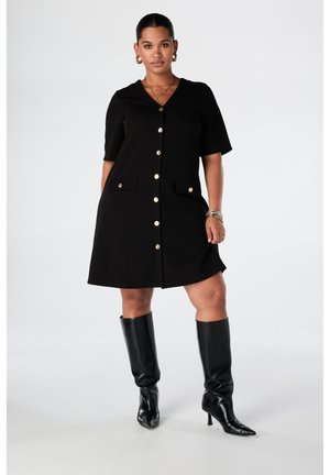 Femme portant une robe noire boutonnée à manches courtes, avec des boutons dorés, deux poches à l'avant, des bijoux en or et des bottes noires à talons montant jusqu'aux genoux.