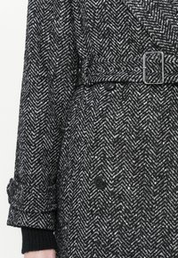 Manteau à motif en chevrons noir et blanc avec une taille ceinturée, featuring des boutons noirs et un tissu texturé. Manches retroussées.