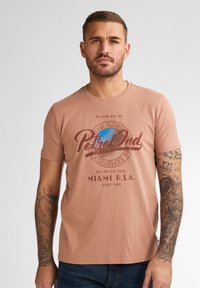 Světle broskvové tričko vyrobené z jemné látky, s kulatým potiskem modrého a hnědého textu a designem glóbusu, nošené s modrými džíny.