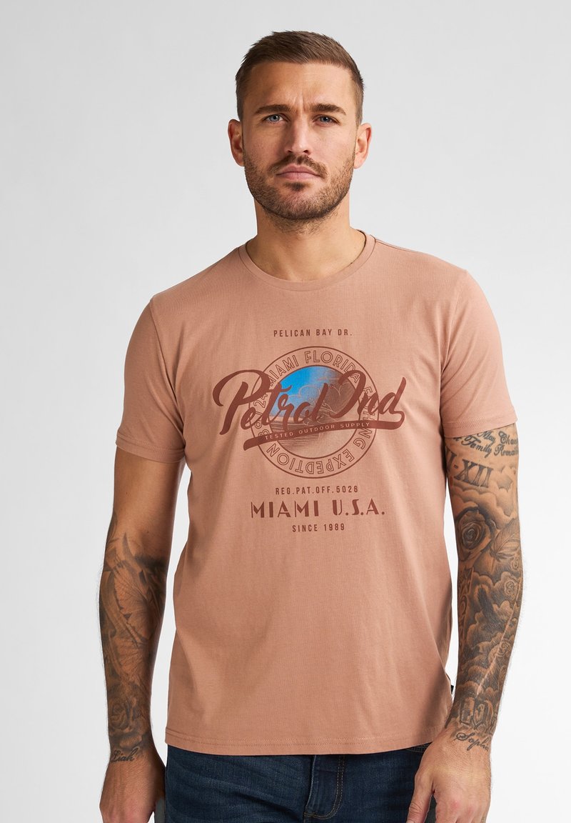 Světle broskvové tričko vyrobené z jemné látky, s kulatým potiskem modrého a hnědého textu a designem glóbusu, nošené s modrými džíny.