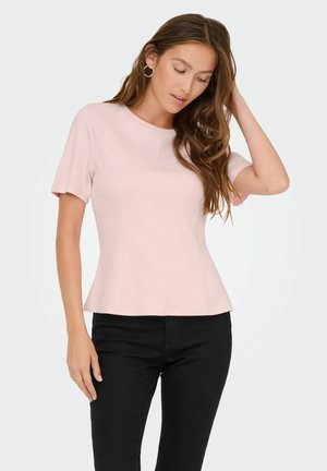 Femme aux longs cheveux bruns portant un haut rose clair à manches courtes et un pantalon noir, touchant ses cheveux et regardant vers le bas.