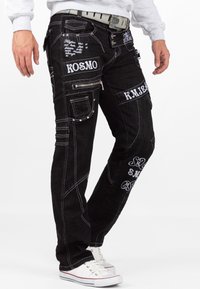 Pantaloni in denim nero con cuciture bianche, multiple tasche, cerniere e testo ricamato. Presentano una vestibilità affusolata e accentuazioni hardware decorative.
