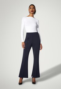 Marineblaue Schlaghosen mit hoher Taille, kombiniert mit einem weißen Langarmoberteil. Das Outfit wird durch schwarze High Heels und eine glatte Textur ergänzt.