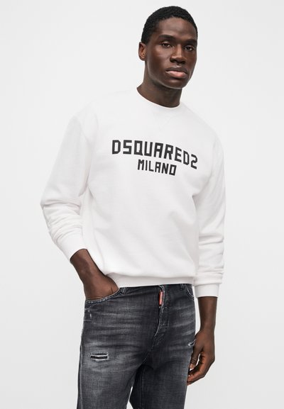 Νέος άνδρας με λευκό φούτερ DSQUARED2 Milano και σκούρα τζιν με ξεβάμματα, ποζάροντας με το ένα χέρι στην τσέπη μπροστά σε λευκό φόντο.