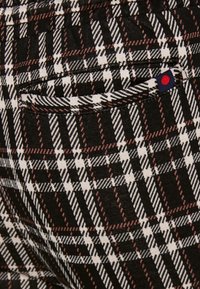 Närbild av svarta, vita och bruna rutiga tygbyxor med en bakficka som har en liten marinblå etikett med en röd prick.