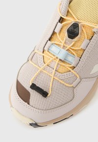 Ανοιχτό γκρι αθλητικό παπούτσι με άνω μέρος από δίχτυ, κίτρινες λεπτομέρειες, ελαστικά κορδόνια και μαύρο κουμπί ρύθμισης. Στρογγυλή μύτη με καουτσούκ σόλα και υφασμένο σχέδιο.