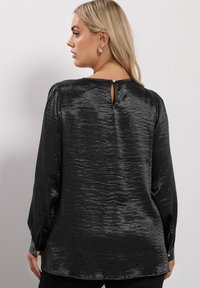 Blusa negra con textura, de mangas largas, con detalle de abertura en la parte posterior y puños abotonados. La tela suave crea un brillo sutil.