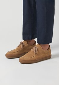 Béžové semišové ležérní boty na šněrování nošené s tmavě modrými zkrácenými kalhotami na jednoduchém světleném pozadí.