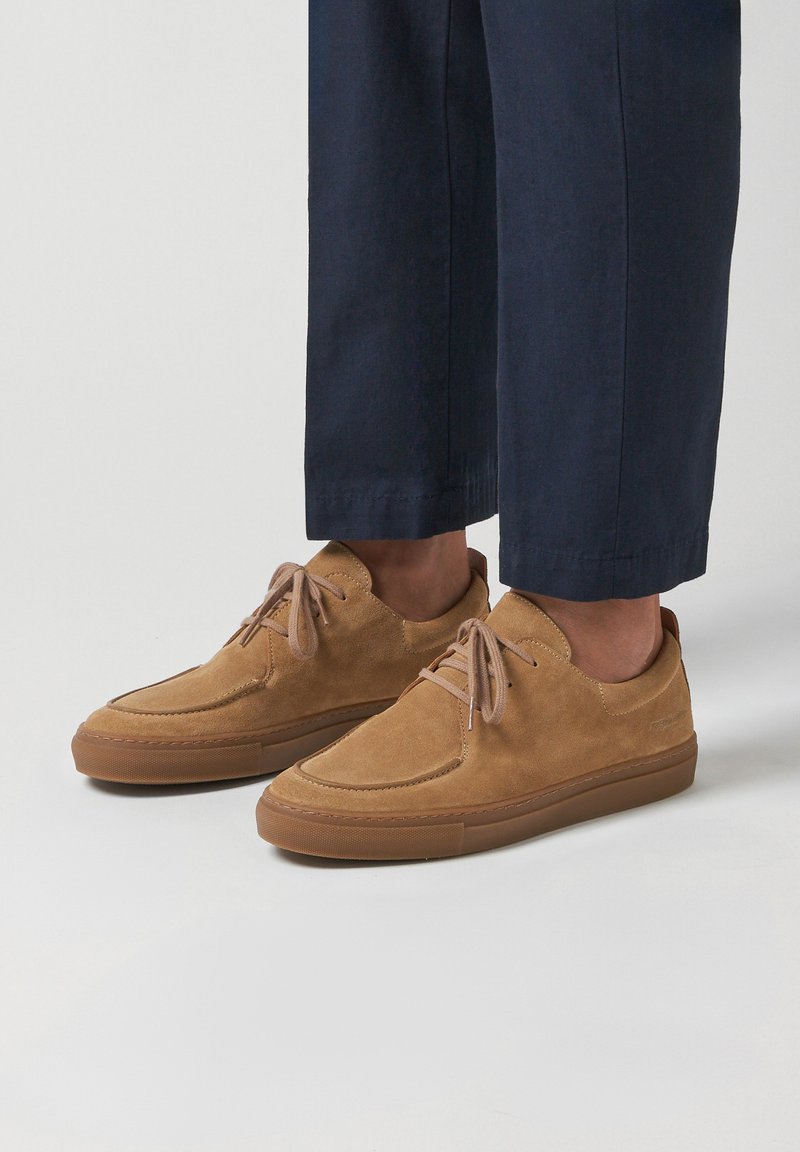 Béžové semišové ležérní boty na šněrování nošené s tmavě modrými zkrácenými kalhotami na jednoduchém světleném pozadí.