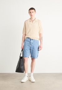 Beige strikket polo skjorte, lyseblå denimshorts, hvite joggesko og en svart tote bag. Korte ermer og en uformell passform er inkludert.