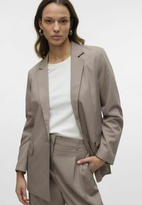 Beige ülikond, mis koosneb sujuvast kangast pintsakust ja sobivatest pükstest, millel on üks nupp, lõikega krae ja küljetaskud.