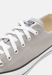 Scarpa da ginnastica bassa grigia in tela con lacci bianchi, occhielli in metallo, doppia cucitura bianca e logo Converse All Star nero e bianco sulla linguetta.