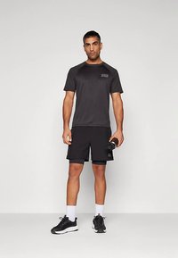 Atletyczny mężczyzna stojący, ubrany w czarny strój sportowy i buty sportowe, trzymający w prawej ręce czarną butelkę na wodę na jednolitym tle.