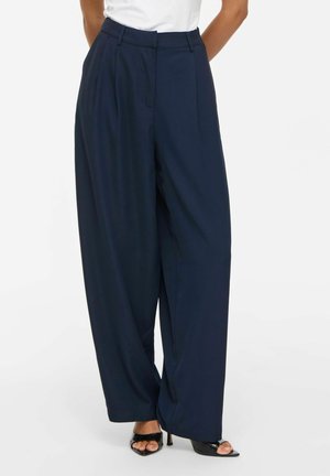 Vrouw die een marineblauwe broek met hoge taille en wijde pijpen draagt, gecombineerd met zwarte open hakken en een wit topje dat is ingestopt.