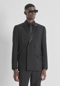 Traje negro cruzado con solapas de satén, combinado con corbata negra y gafas de sol negras. La tela parece suave y de corte entallado.