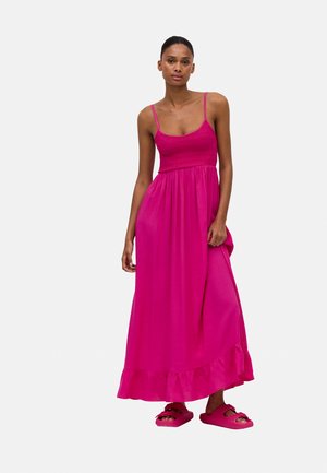 Vestido maxi fucsia con un torso ajustado, textura fruncida y dobladillo con volantes. Lleva unos sandalias de dedo rosas con tiras anchas.