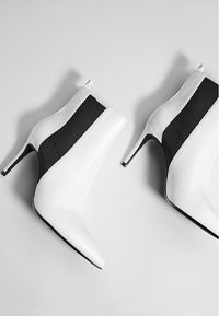 Botines blancos hasta el tobillo con punta afilada, que presentan un panel elástico negro en los laterales. Tacón de aguja alto con textura suave.