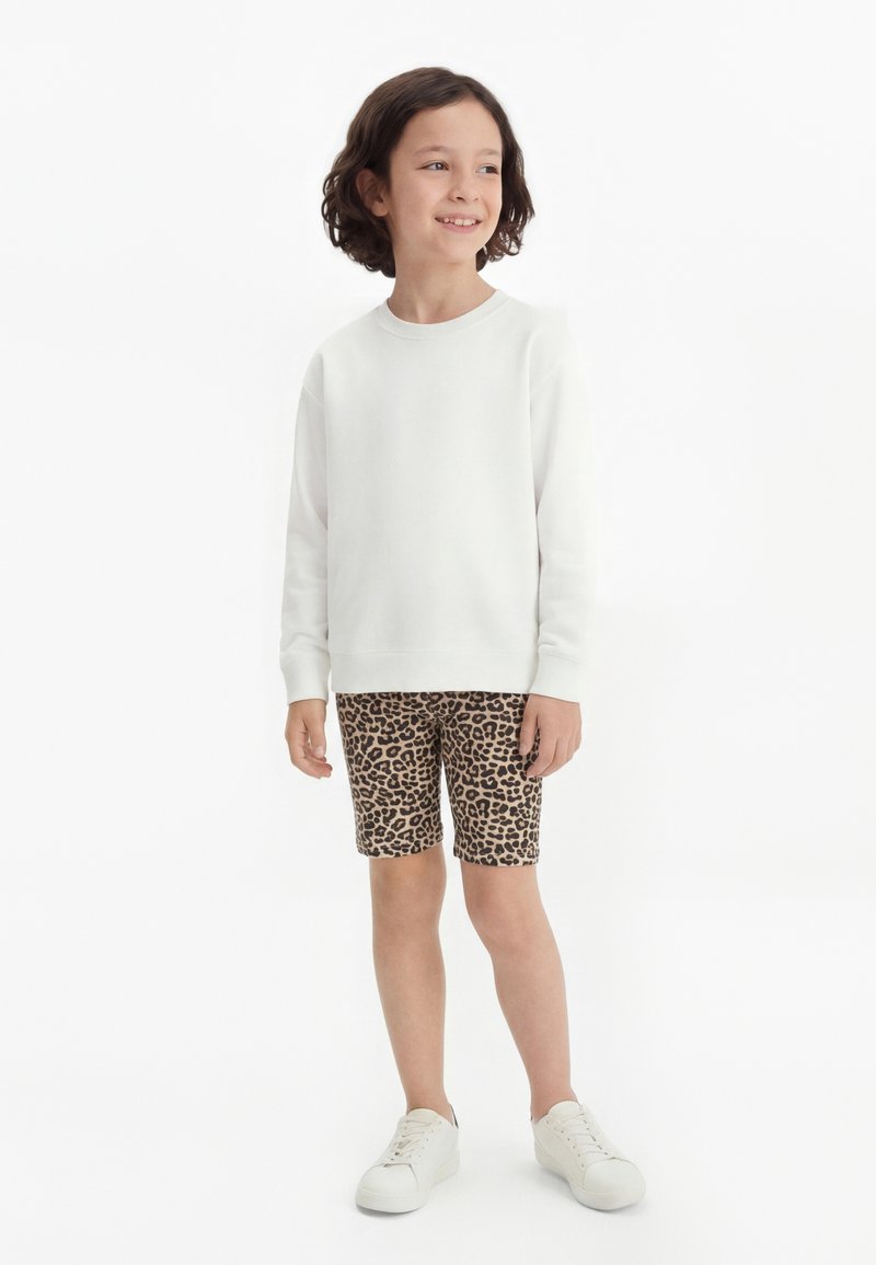 Enfant aux cheveux courts et bouclés, debout en souriant, portant un t-shirt blanc à manches longues, un short imprimé léopard et des baskets blanches sur fond blanc.