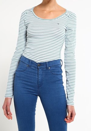 Femme portant un haut à manches longues blanc et vert à rayures horizontales avec un col en U et un jean bleu taille haute ajusté.