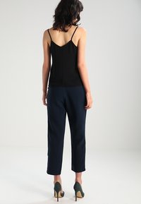 Czarna koszulka na ramiączkach z cienkimi paskami, zestawiona z granatowymi spodniami w stylu tapered. Modelka nosi zielone szpilki; widoczne są gładki materiał i proste linie.