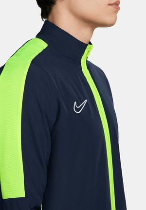 Marineblaue Sportjacke mit neongrünen Mesh-Einsätzen und Reißverschluss. Sie verfügt über einen hohen Kragen und ein gesticktes weißes Logo auf der Brust.