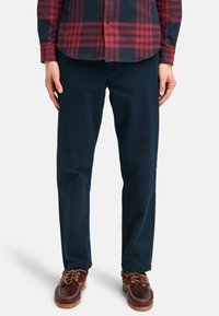 Navy blue cotton chinos with a straight leg cut, paired with a red and black plaid shirt. Brown leather shoes with white stitching.
