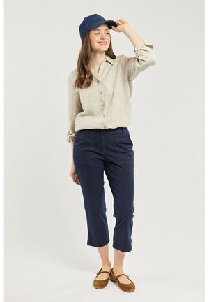 Femme souriante ajuste une casquette bleue portant une chemise beige boutonnée, un pantalon court marine et des chaussures plates marron, debout sur un fond neutre.