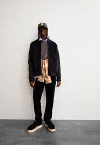 Jeune homme portant une veste noire sur un pull marron, une chemise à carreaux jaune nouée autour de la taille, un pantalon noir, des chaussures à plateforme et une casquette grise.