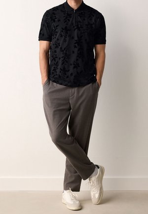Homme portant un polo noir à manches courtes avec motif floral, pantalon marron, baskets blanches, debout les mains dans les poches et les jambes croisées.