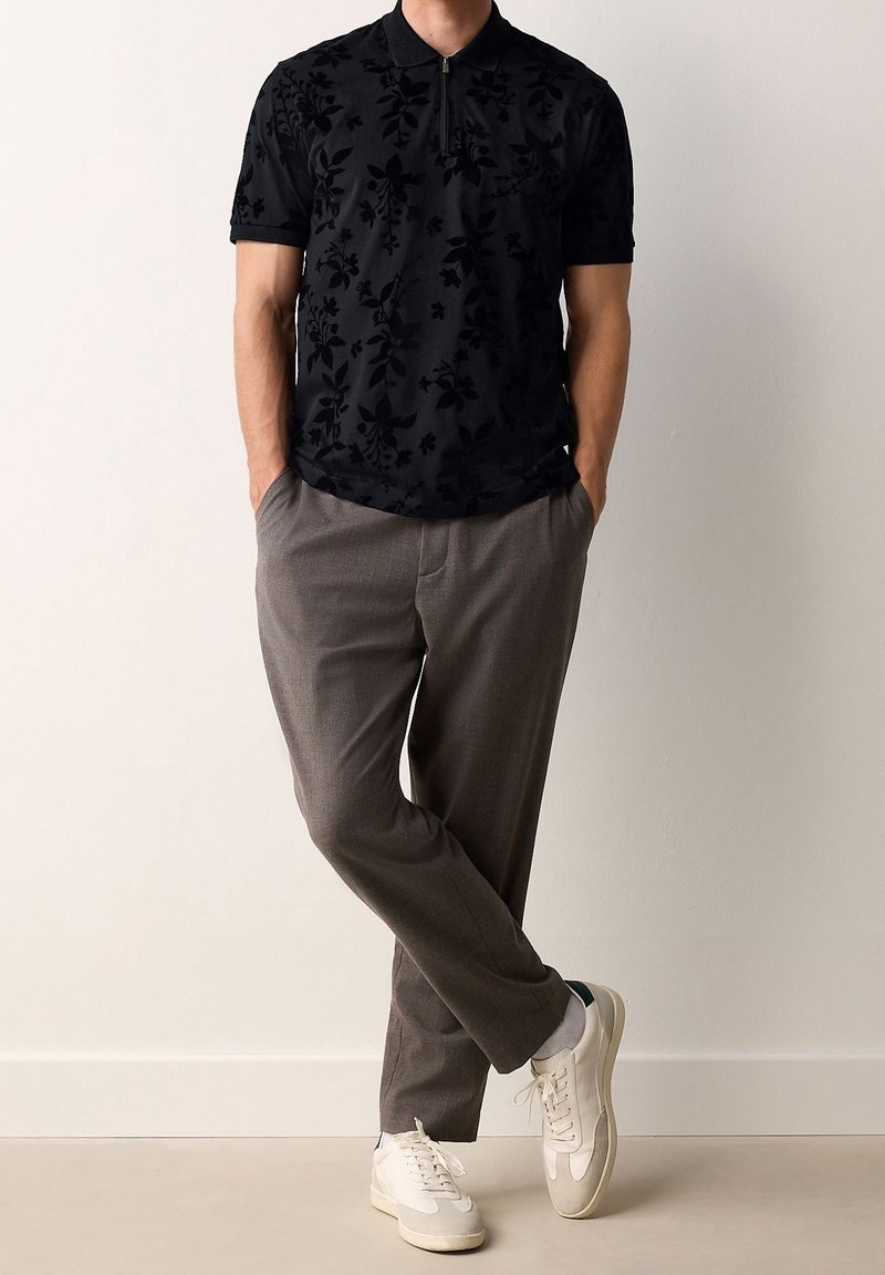 Homme portant un polo noir à manches courtes avec motif floral, pantalon marron, baskets blanches, debout les mains dans les poches et les jambes croisées.