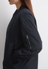 Veste d'aviateur bleu marine avec une texture lisse et légère, des poignets côtelés et une poche zippée sur la manche. Design minimaliste et lignes épurées.
