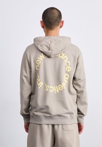 Beige kapucnis pulóver, amelyen a hátsó részen egy kör alakú sárga grafikus szöveg található, puha tapintású, bordás mandzsettákkal és aljjal.