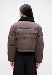 Brązowa pikowana kurtka z czarnym kołnierzem z owczej wełny, z wytłoczonym napisem "Tommy Jeans" z tyłu. Krótki krój, gładka faktura.