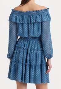 Kvinna som bär en blå off-shoulder klänning med vita prickar, långa genomskinliga ärmar, volangskikt och elasticiserad halsringning och manschetter.