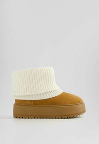 Bottine en daim marron avec un large revers en maille côtelée crème et une semelle plateforme robuste couleur tabac. Surface texturée et contraste de couleurs distinct.