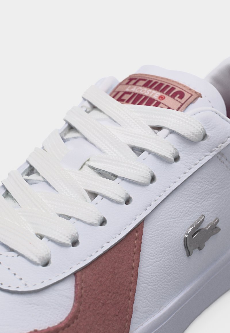 White leather sneaker with pink suede detail, white laces, metal crocodile logo, and branded tag on tongue.