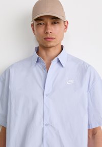 Lys blå kortermet skjorte med knapper og avslappet passform, med en hvit Nike-logo på venstre bryst og et strukturert stoff.