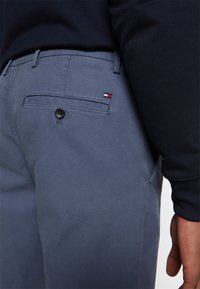 Homme portant un pantalon bleu texturé avec une poche arrière à bouton et un petit logo rouge, blanc et bleu, assorti à une chemise noire à manches longues.