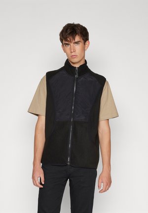 Gilet en polaire noire avec fermeture éclair sur le devant et col haut. Panneau en tissu sombre sur la poitrine, présentant des textures et des détails de couture contrastants.